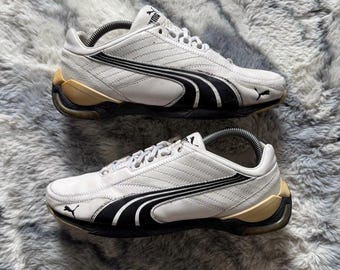 May include: Pair of white Puma sneakers with black and gold accents. The shoes feature the Puma logo and a streamlined design, suitable for athletic activities or casual wear.