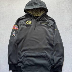 May include: A dark grey hooded sweatshirt featuring the Green Bay Packers logo, a Salute to Service patch, an American flag patch, and an NFL ribbon patch. The hood interior has a camouflage pattern, and a small yellow Nike swoosh is on the sleeve.