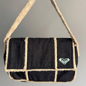 Puede incluir: Bolso de hombro de mezclilla negra con ribete y correa de color crema y esponjoso. El bolso tiene forma cuadrada con solapa y un pequeño logotipo turquesa. El bolso está hecho de un material suave y texturizado.