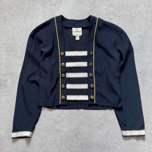 Peut inclure: Une veste bleu marine à manches longues avec un panneau central composé de cinq bandes horizontales blanches bordées d'or. La veste présente un col en V, des boutons dorés sur les bords avant et des poignets rayés blanc et or. L'étiquette Joseph Ribkoff est visible.