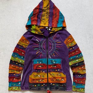 Puede incluir: Una sudadera con capucha y cremallera de color morado, con capucha puntiaguda, bordado central de sol y paneles de patchwork horizontales en mangas y bolsillos delanteros. La prenda presenta un diseño multicolor con tiras de tela roja, amarilla, turquesa y negra.