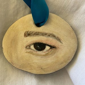 May include: Oval-shaped pendant featuring a painted eye with a dark iris, light skin tone, and a brown eyebrow. The pendant has a cracked, aged appearance and is suspended by a teal ribbon.