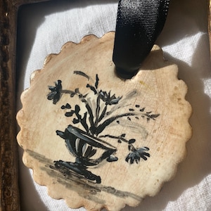 May include: A round, off-white ornament with a scalloped edge, featuring a black and gray floral design in a vase. A black ribbon is attached at the top. The ornament is set within a gold-colored frame.