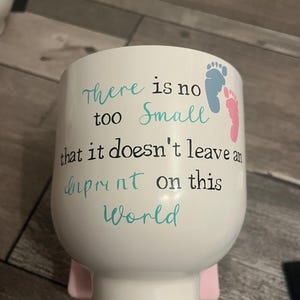 Könnte beinhalten: Weißer Keramikbecher mit rosa Sockel, mit dem Text "there is no too small that it doesn't leave an imprint on this world." Das Design umfasst blaue und rosa Fußabdrücke.