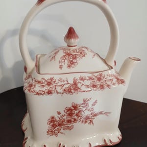 May include: Cream-colored ceramic teapot with a red floral pattern. The teapot has a curved handle, a lid with a conical finial, and a spout. The floral design decorates the body and lid. The base has a scalloped edge.