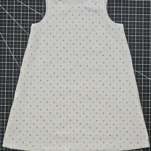May include: A sleeveless white dress with a pattern of small, evenly spaced dots in pastel colors. The dress has a rounded neckline and two small buttons on each shoulder. The fabric appears to be lightweight, suitable for warm weather.