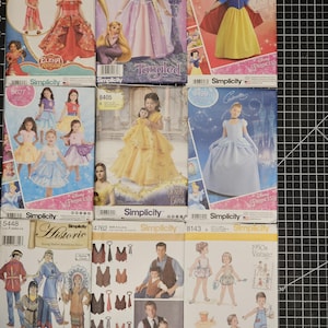 May include: A collection of nine sewing patterns for children's clothing. The patterns feature various Disney characters, including princesses like Cinderella, Snow White, and Belle, as well as other characters like Tangled's Rapunzel and Elena of Avalor. The patterns are from Simplicity and include instructions for making dresses, shirts, and other garments.