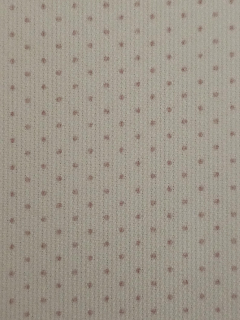 Op de afbeelding: Een close-up van een witte stof met een subtiel roze stippenpatroon. De stof heeft een textuur, een licht bobbelige oppervlak.