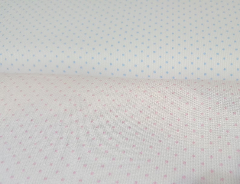 Op de afbeelding: Twee stukken stof met een witte achtergrond en kleine, gelijkmatig verdeelde stippen. Het bovenste stuk stof heeft lichtblauwe stippen, en het onderste stuk stof heeft roze stippen.