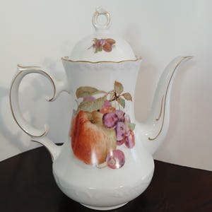 May include: White porcelain coffee pot with a lid, handle, and spout. The pot is decorated with a colorful fruit design featuring an apple, cherries, and other fruits. Gold trim accents the edges of the pot and lid.