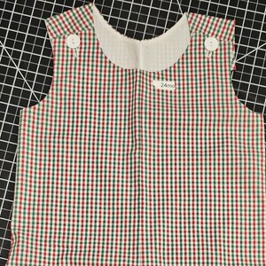 Puede incluir: Un pelele de bebé sin mangas con un estampado de cuadros rojos, verdes y blancos. El pelele tiene un cuello blanco y dos botones blancos en cada hombro. La etiqueta dice "24mo".