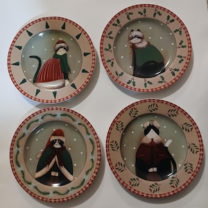 May include: Set of four decorative plates featuring whimsical cat illustrations in winter attire. Each plate has a beige border with a red and green festive design. The cats are depicted in various outfits, with a snowy background.
