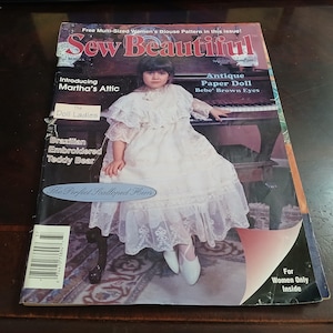 May include: A vintage magazine cover titled "Sew Beautiful" features a young girl in a white dress. The cover includes text such as "Free Multi-Sized Women's Blouse Pattern" and "Antique Paper Doll." The magazine is on a dark wooden surface.