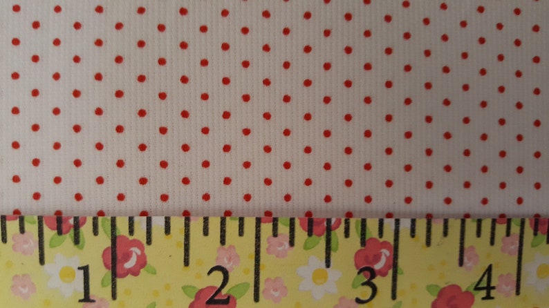 Op de afbeelding: Witte stof met een rood stippenpatroon. Een liniaal met bloemmotieven is onderaan de afbeelding geplaatst. De liniaal toont metingen van 1 tot 4 inch.
