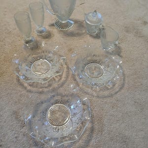 May include: A clear glass serving set featuring a pitcher, two stemmed glasses, a sugar bowl with a lid, a creamer, and three decorative bowls. The glassware has a floral design and a scalloped edge.