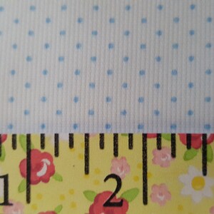 Op de afbeelding: Een close-up van een witte stof met een lichtblauw stippenpatroon. Een gele meetlint met zwarte markeringen en een bloemenprint bevindt zich onderaan de afbeelding. De lint geeft "1", "2" en "3" aan.