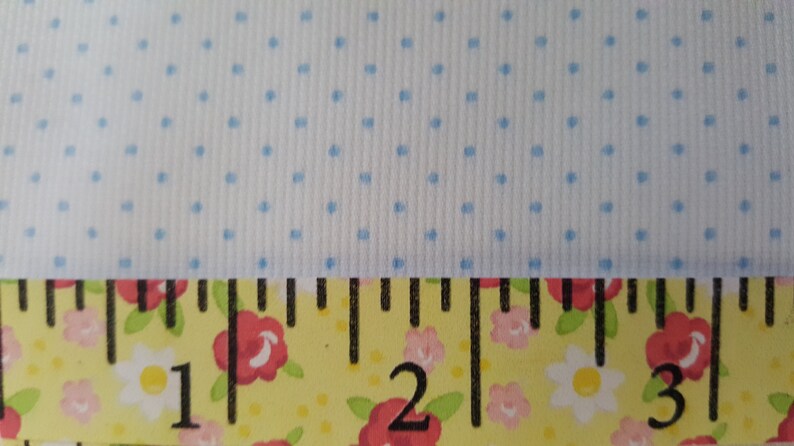 Op de afbeelding: Een close-up van een witte stof met een lichtblauw stippenpatroon. Een gele meetlint met zwarte markeringen en een bloemenprint bevindt zich onderaan de afbeelding. De lint geeft "1", "2" en "3" aan.