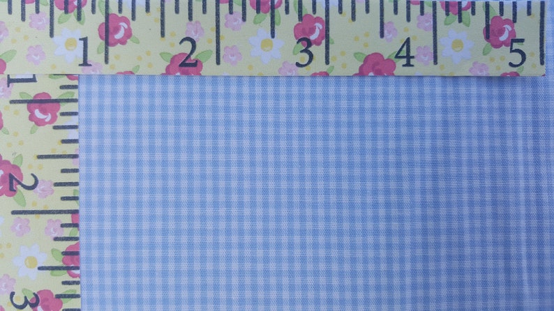 K&ouml;nnte beinhalten: Nahaufnahme eines hellblauen Gingham-Stoffes mit einem wei&szlig;en Gittermuster. Oben im Bild ist ein Lineal mit floralen Akzenten zu sehen.