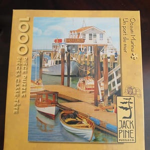 May include: A 1000-piece jigsaw puzzle featuring a harbor scene with boats docked at a wooden pier. The puzzle box displays the words "Ocean Marina" and "Danfords". The Jack Pine Puzzle Co. logo is visible on the box.