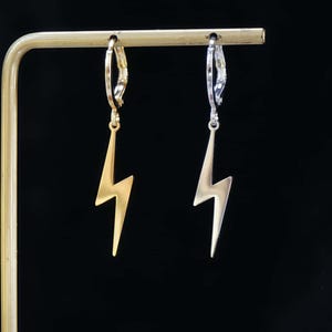 May include: A pair of gold and silver-colored lightning bolt earrings hanging from a gold-colored display stand. The earrings have a simple, minimalist design, with a hook closure.