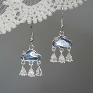 May include: Silver-tone cloud earrings with blue enamel and dangling teardrop-shaped clear crystals. Each cloud has a small star accent. The earrings hang from silver-tone fish hook ear wires. The earrings are a whimsical accessory.
