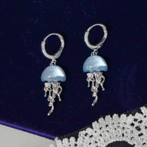 May include: A pair of silver-toned jellyfish earrings. Each earring features a light blue, dome-shaped top and intricate, dangling tendrils. The earrings are set against a dark blue background.
