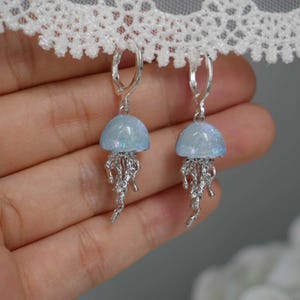 May include: A pair of earrings featuring a jellyfish design. Each earring has a light blue, dome-shaped top and silver-colored, dangling tentacles. The earrings are suspended from silver-colored lever-back closures. The background is blurred.