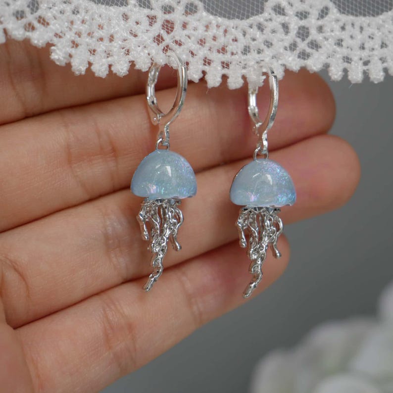 May include: A pair of earrings featuring a jellyfish design. Each earring has a light blue, dome-shaped top and silver-colored, dangling tentacles. The earrings are suspended from silver-colored lever-back closures. The background is blurred.