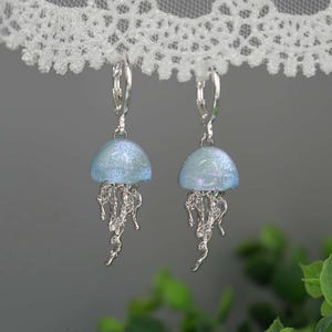 May include: A pair of silver-toned jellyfish earrings. Each earring features a light blue, dome-shaped top with iridescent glitter and dangling, detailed tentacles. The earrings hang from silver lever-back closures.