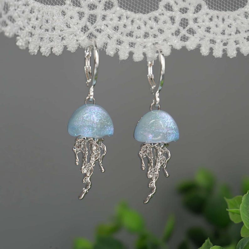 May include: A pair of silver-toned jellyfish earrings. Each earring features a light blue, dome-shaped top with iridescent glitter and dangling, detailed tentacles. The earrings hang from silver lever-back closures.
