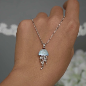 May include: A silver-toned necklace featuring a pendant shaped like a jellyfish. The jellyfish pendant has a light blue, glittery top and detailed silver tentacles. The necklace is displayed against a neutral background.