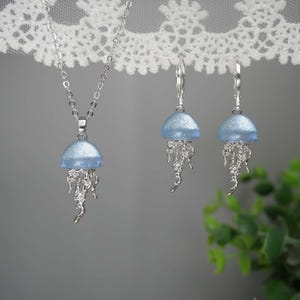 May include: A silver necklace and earrings set featuring a jellyfish design. The pendant and earrings have a light blue, dome-shaped top and silver, flowing tentacles. The jewelry is displayed against a soft gray background.