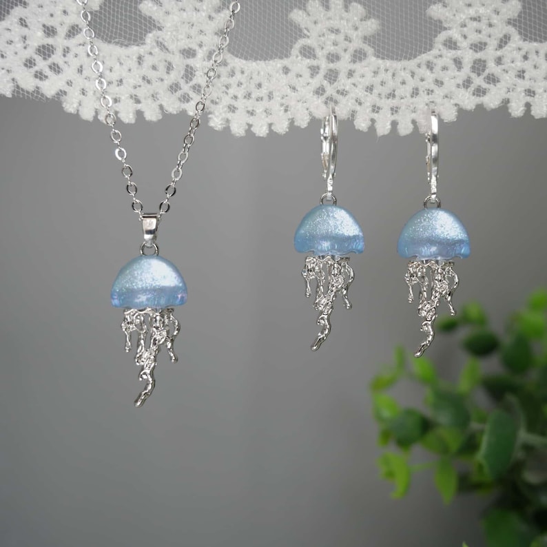 May include: A silver necklace and earrings set featuring a jellyfish design. The pendant and earrings have a light blue, dome-shaped top and silver, flowing tentacles. The jewelry is displayed against a soft gray background.