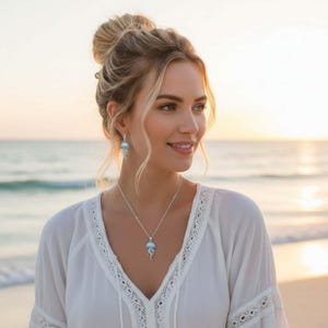 May include: A woman wearing a silver necklace and earrings with a jellyfish design. The jewelry has a light blue stone and a detailed silver design. The woman is wearing a white top and is standing on a beach.
