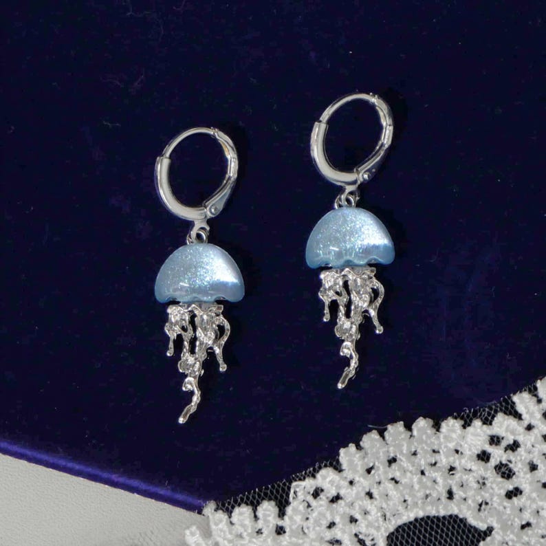 May include: A pair of silver-toned jellyfish earrings. Each earring features a light blue, dome-shaped top and intricate, dangling tendrils. The earrings are set against a dark blue background.