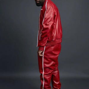 May include: Red leather track suit with white stripes. The jacket has a ribbed collar and cuffs. The pants have a straight leg design. The outfit is paired with white sneakers. The background is a solid gray.