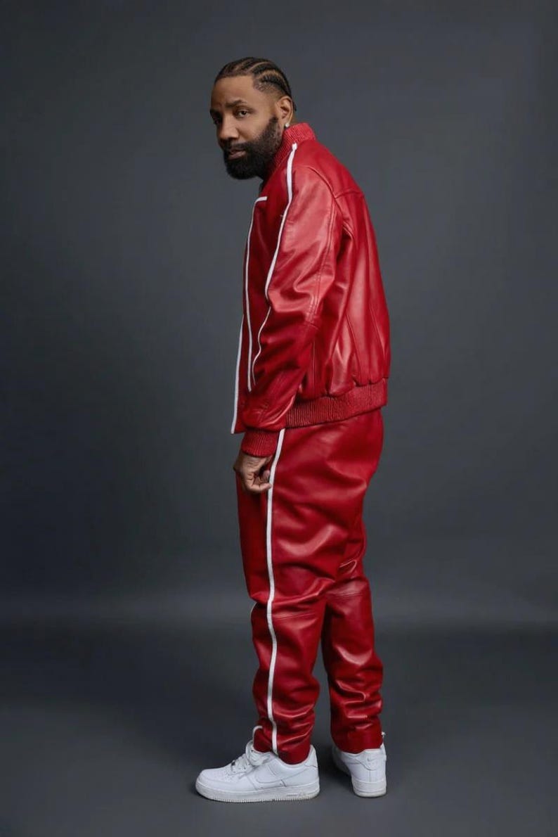 May include: Red leather track suit with white stripes. The jacket has a ribbed collar and cuffs. The pants have a straight leg design. The outfit is paired with white sneakers. The background is a solid gray.