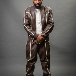 May include: A brown leather tracksuit with white trim. The jacket has a bomber style with a ribbed collar and cuffs. The pants are straight-legged. The outfit is paired with white sneakers. The person is wearing a white t-shirt underneath.
