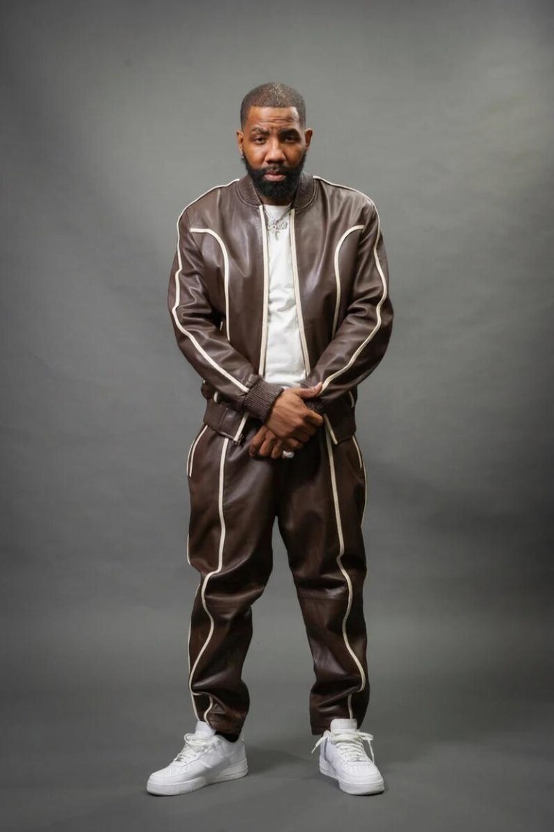 May include: A brown leather tracksuit with white trim. The jacket has a bomber style with a ribbed collar and cuffs. The pants are straight-legged. The outfit is paired with white sneakers. The person is wearing a white t-shirt underneath.