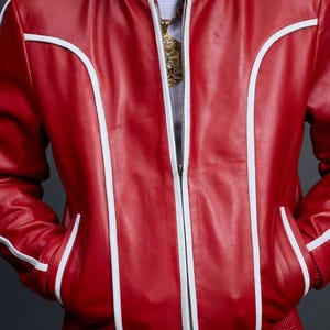 May include: A red leather bomber jacket with white trim accents. The jacket features a zipper closure, pockets, and a ribbed collar and cuffs. The jacket is part of a matching set.