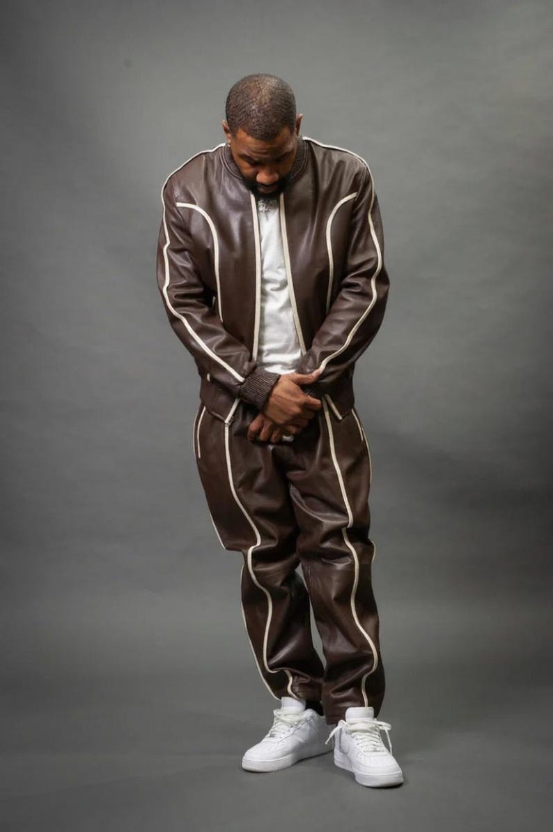 May include: Brown leather tracksuit with white piping. The jacket has a bomber style with a white t-shirt underneath. The matching pants have a relaxed fit. The outfit is paired with white sneakers. The model is standing in front of a gray background.