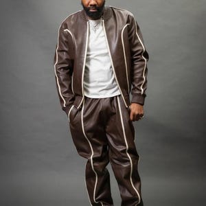 May include: A brown leather tracksuit with white trim. The jacket has a zip-up front and the pants have pockets. The outfit is paired with white sneakers. The person is wearing a white t-shirt underneath the jacket.