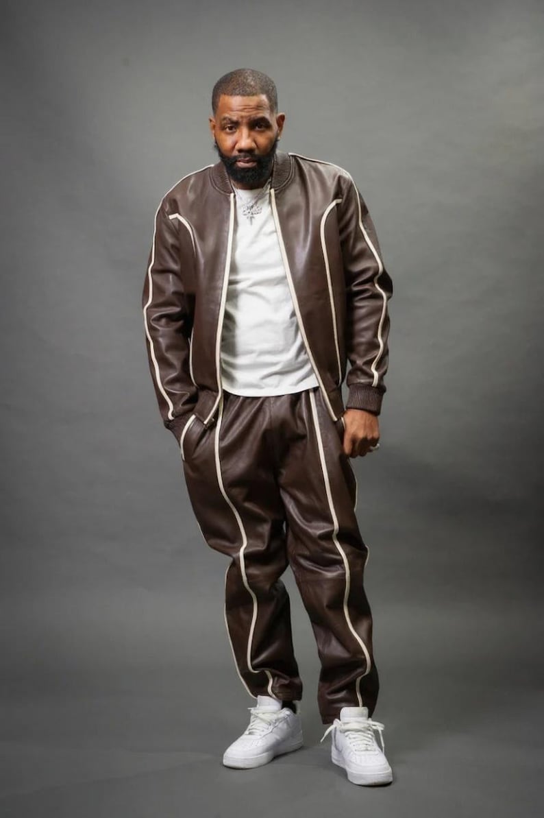 May include: A brown leather tracksuit with white trim. The jacket has a zip-up front and the pants have pockets. The outfit is paired with white sneakers. The person is wearing a white t-shirt underneath the jacket.