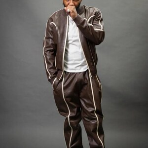 May include: A brown leather tracksuit with white trim, consisting of a bomber jacket and matching pants. The outfit is paired with a white t-shirt and white sneakers. The person is standing in front of a gray background.