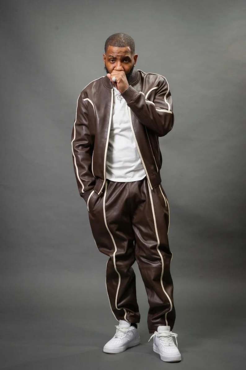 May include: A brown leather tracksuit with white trim, consisting of a bomber jacket and matching pants. The outfit is paired with a white t-shirt and white sneakers. The person is standing in front of a gray background.