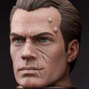 May include: A sculpted bust of a man's head, with short, dark brown hair and a serious expression. The face displays realistic skin tones, with scars and fine details. The eyes are a vivid green. The bust is presented against a dark backdrop.