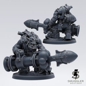 Könnte beinhalten: Zwei graue Miniatur-Orc-Figuren, eine vorne und eine hinten, die jeweils eine große, stilisierte Kanone halten. Die Figuren sind detailliert mit Rüstung und Gesichtszügen. Das Smuggler Boutique-Logo befindet sich unten rechts.