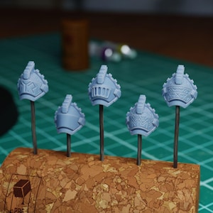 May include: Five light blue miniature helmets with intricate designs are displayed on metal rods inserted into a corkboard. The helmets have a detailed, textured appearance, suggesting they are part of a model or miniature set.