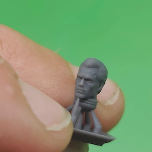 May include: A small, grey miniature head with detailed facial features, including a furrowed brow and defined jawline. The head is attached to a base with supporting structures, likely for a miniature figure or model. The background is a solid green.