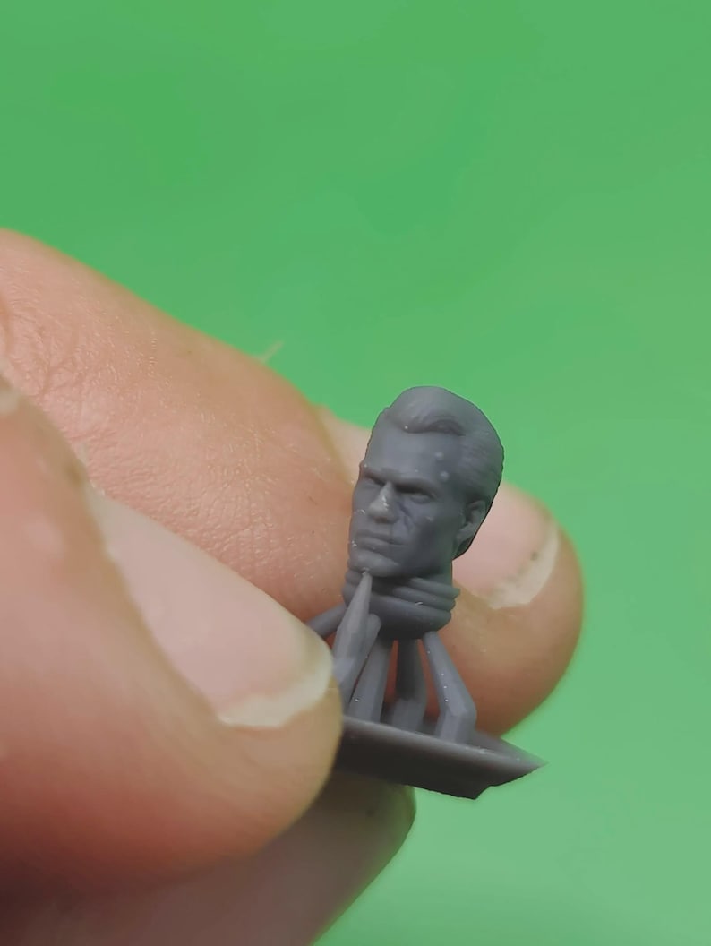 May include: A small, grey miniature head with detailed facial features, including a furrowed brow and defined jawline. The head is attached to a base with supporting structures, likely for a miniature figure or model. The background is a solid green.